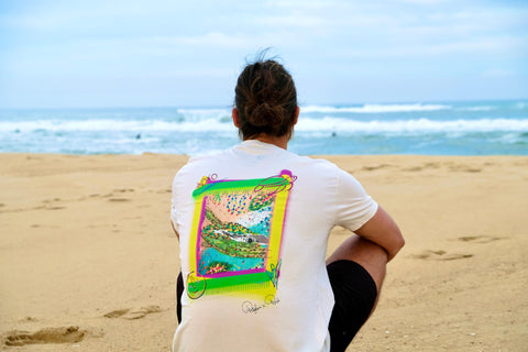 Mann sitzt am Strand und schaut auf den Ozean, er trägt ein weißes Bio-T-Shirt mit Schildkrötenaufdruck